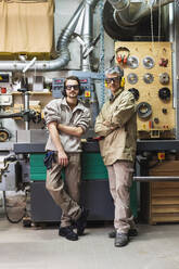 Male colleagues wearing ear protectors and protective eyewear standing by machine - MEUF03605