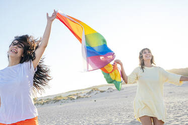 Cheerful young women holding rainbow flag while waking at beach - JCMF02116