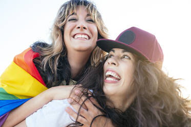 Cheerful woman with rainbow flag piggybacking on girlfriend - JCMF02126