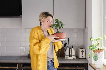 Woman kissing houseplant in kitchen at home - KMKF01718
