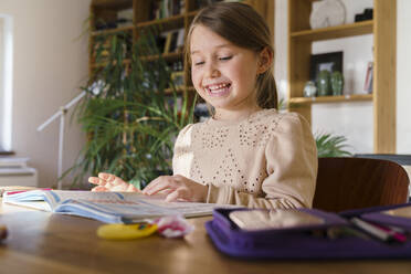 Smiling girl looking at book on table while studying at home - KMKF01721