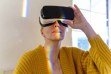 Woman with spectrum on face wearing virtual reality headset at home - KMKF01725