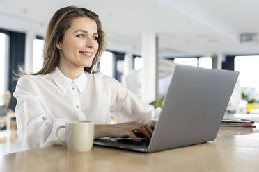 Smiling businesswoman using laptop on table at office - PESF02956