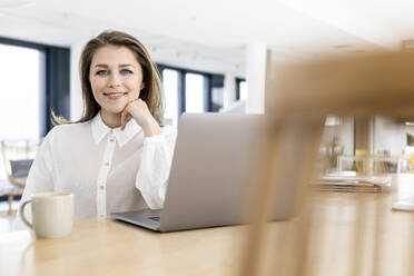 Businesswoman with hand on chin sitting by laptop at office - PESF02957