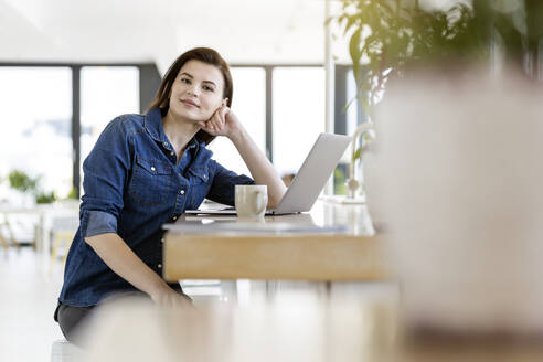 Geschäftsfrau lehnt sich mit ihrem Laptop im Büro an einen Tisch - PESF02971