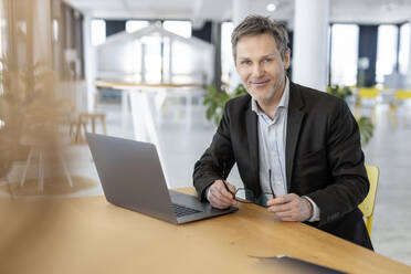 Smiling businessman with laptop sitting at office - PESF03015