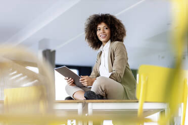 Afro businesswoman holding digital tablet while sitting with cross-legged at office - PESF03029