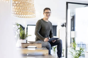 Smiling mature businessman sitting with digital tablet on desk in office - PESF03055