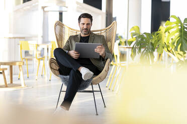 Männlicher Fachmann, der ein digitales Tablet benutzt, während er auf einem Korbstuhl im Büro sitzt - PESF03078