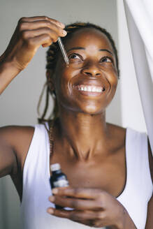 Lächelnde Afro-Frau, die beim Auftragen von Gesichtsserum nach oben schaut - OCMF02191