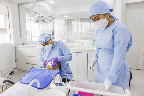 Stomatologin, die die Zähne eines nicht erkennbaren männlichen Patienten behandelt, gegen einen Mitarbeiter in Uniform im Krankenhaus - ADSF27104
