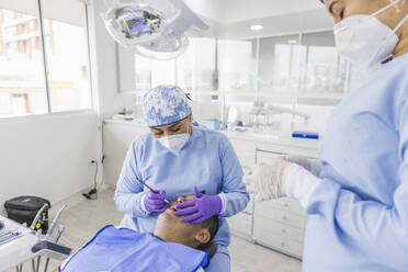 Female stomatologist in uniform and respiratory mask curing teeth of male patient in hospital - ADSF27105