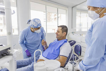 Smiling man looking in mirror while talking to female doctors in uniforms after dental procedure in clinic - ADSF27106