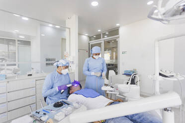 Eine Ärztin in medizinischer Uniform und mit Maske heilt Zähne mit einem UV-Gerät, während sie mit einem Kollegen in einer modernen Zahnklinik arbeitet - ADSF27113