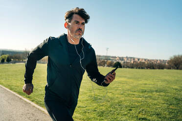 Erwachsener männlicher Jogger in Sportkleidung, der auf dem Bürgersteig zwischen Rasenflächen läuft, während er beim Training in der Stadt nach vorne schaut - ADSF27123