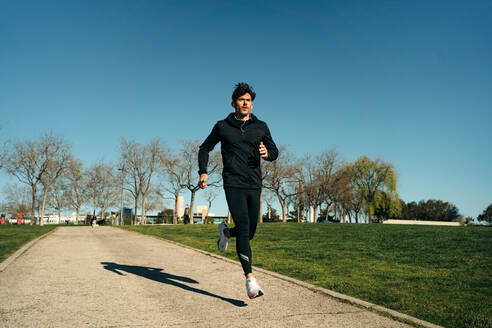 Erwachsener männlicher Jogger in Sportkleidung, der auf dem Bürgersteig zwischen Rasenflächen läuft, während er beim Training in der Stadt nach vorne schaut - ADSF27124