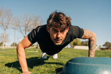 Ground level of determined male athlete in sportswear training on meadow while looking forward under blue sky - ADSF27127