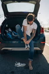 Tätowierter männlicher Sportler, der sich Schuhe anzieht, während er mit einem Mobiltelefon gegen ein Auto mit offenem Kofferraum in der Stadt spricht - ADSF27129