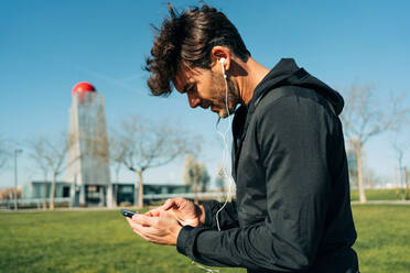 Bärtiger männlicher Sportler in Sportkleidung und Kopfhörern, der auf einer Wiese in der Stadt steht und mit seinem Handy SMS schreibt - ADSF27130
