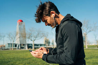 Bearded male athlete in sportswear and earphones text messaging on cellphone while standing on meadow in town - ADSF27130