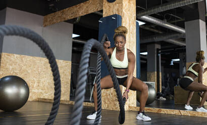 Starke afroamerikanische Sportlerin in Sportkleidung, die mit Kampfseilen trainiert, während sie bei einem hochintensiven Training im Fitnessstudio nach vorne schaut - ADSF27136