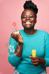 Happy African American female with eyes closed wearing blue clothes and blowing soap bubbles against pink background in studio - ADSF27167