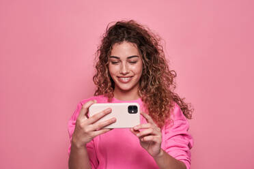 Smiling teenage with curly hair while taking self portrait on mobile phone against pink background - ADSF27176