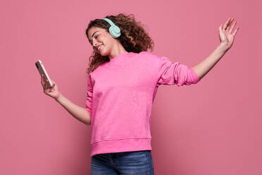 Cheerful teenager with curly hair outstretching arm and scrolling mobile phone while listening to music and jumping against pink background - ADSF27177