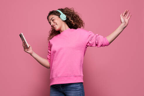 Fröhlicher Teenager mit lockigem Haar, der den Arm ausstreckt und auf seinem Handy scrollt, während er Musik hört und vor einem rosa Hintergrund springt - ADSF27177