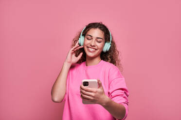 Cheerful teenager with curly hair scrolling mobile phone while listening to music standing against pink background - ADSF27178