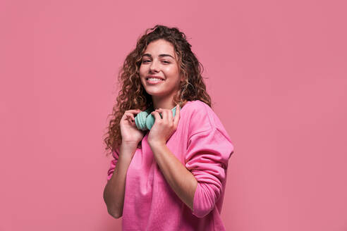 Glückliche Teenagerin mit lockigem Haar in Freizeitkleidung, die in die Kamera schaut, mit drahtlosen Kopfhörern auf rosa Hintergrund - ADSF27179