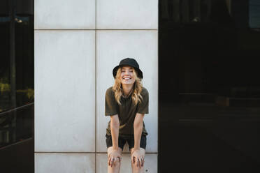 Lächelnde junge Frau in Freizeitkleidung, die sich nach vorne lehnt und in die Kamera schaut, in einer Stadt bei Tageslicht - ADSF27189