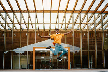 Low angle full body of African American male with headphones on neck jumping high with arms outstretched - ADSF27198