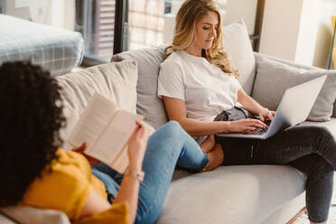 Lesbisches Paar, das auf einem Netbook surft und ein interessantes Buch liest, während es sich auf einer bequemen Couch im modernen Wohnzimmer ausruht - ADSF27205