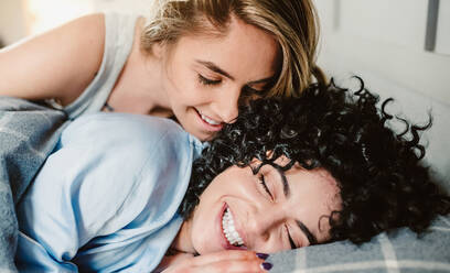 Cheerful crop girlfriend awakening curly haired lesbian partner lying in bed with closed eyes in morning - ADSF27212