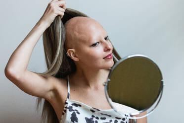 Junge selbstbewusste Frau mit Alopezie, die in den Spiegel schaut und ihre Perücke abnimmt, vor weißem Hintergrund - ADSF27234