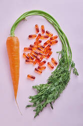 Von der obigen Zusammensetzung der verstreuten Vitaminpillen, die auf einem rosa Tisch in der Nähe von reifen Karotten angeordnet sind - ADSF27249