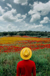 Rückenansicht unerkennbar stilvolle männliche in roter Kleidung und gelben Hut stehend in üppigen blühenden Feld in friedlicher Natur - ADSF27256