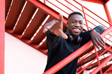 Delighted young African American male with headphones on neck raising arm trying to reach gesture and looking at camera while standing on outdoor metal staircase - ADSF27257