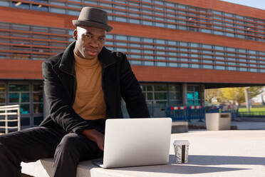 Nachdenklicher afroamerikanischer Mann in stilvollem Outfit, der ein Netbook benutzt und mit einem Kaffee zum Mitnehmen auf einer sonnigen Straße sitzt und in die Kamera schaut - ADSF27262