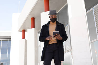 Low angle positive African American male in stylish wear and face mask browsing tablet while standing on sunny street - ADSF27263