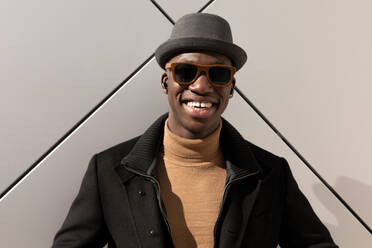 Trendiger, selbstbewusster, fröhlicher Afroamerikaner mit Hut und Sonnenbrille, der an einer grauen Wand steht und in die Kamera schaut - ADSF27265