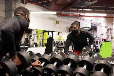 Selbstbewusster junger afroamerikanischer Sportler in Sportkleidung und Maske hebt eine schwere Hantel, während er auf einer Bank in einem modernen Fitnessstudio sitzt - ADSF27272