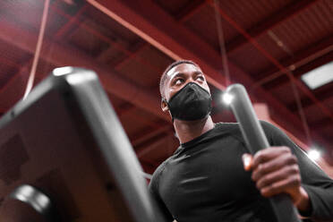 From below African American sportsman in mask doing cardio workout on elliptical machine in modern gym - ADSF27273
