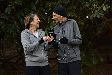 Smiling senior couple in sportswear and earphones standing together under tree branches and sharing mobile phone during fitness workout - ADSF27275