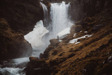 Blick in die Ferne auf einen Touristen, der auf einem hohen Felsen vor einer fließenden Wasserfallkaskade im Hintergrund steht, Island - ADSF27281