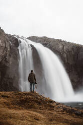 Rückansicht eines Mannes mit Fotokamera, der auf einem Felsen vor einem beeindruckenden Wasserfall auf einer Klippe steht, Island - ADSF27286