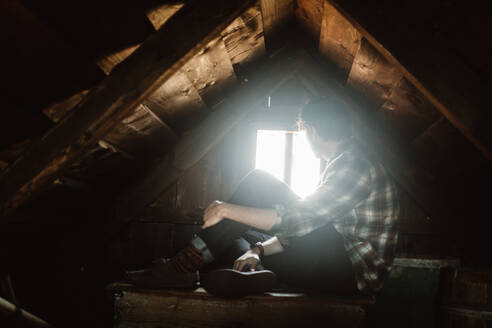 Seitenansicht eines nicht erkennbaren jungen Mannes, der auf einem dunklen Dachboden sitzt und aus einem kleinen Fenster in einer isländischen Landschaft schaut - ADSF27287