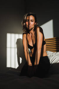 Aufnahme einer sinnlichen hübschen Frau, die zwischen Licht und Schatten lächelt, in Dessous auf dem Bett und in die Kamera schaut - ADSF27303
