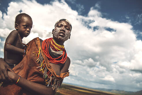 Young woman with painted face holding her little boy, against cloudy sky, in Karo tribe village. Omo Valley, Ethiopia - ADSF27322